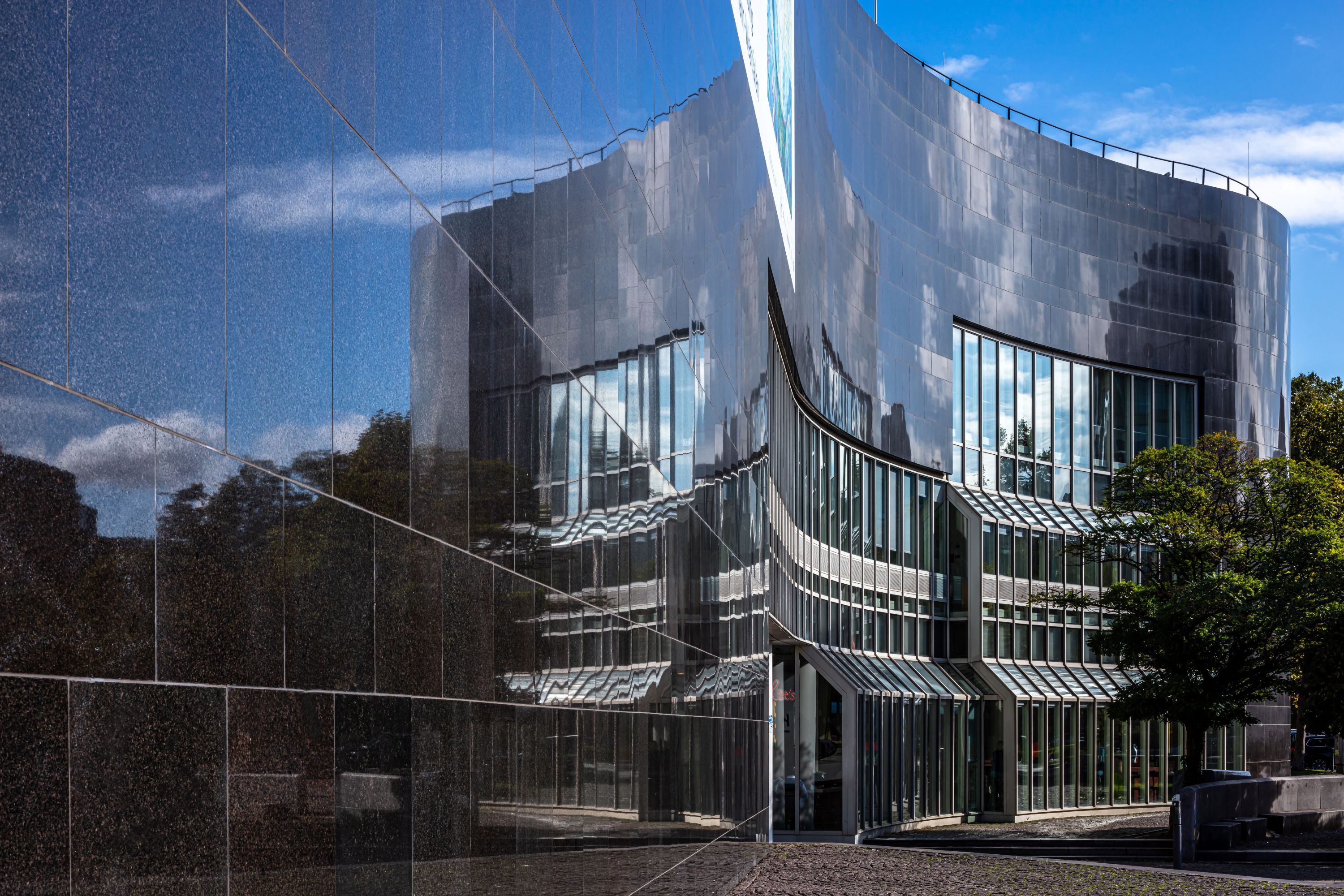 Geschwungene Fassade, in der sich Wolken und Bäume spiegeln